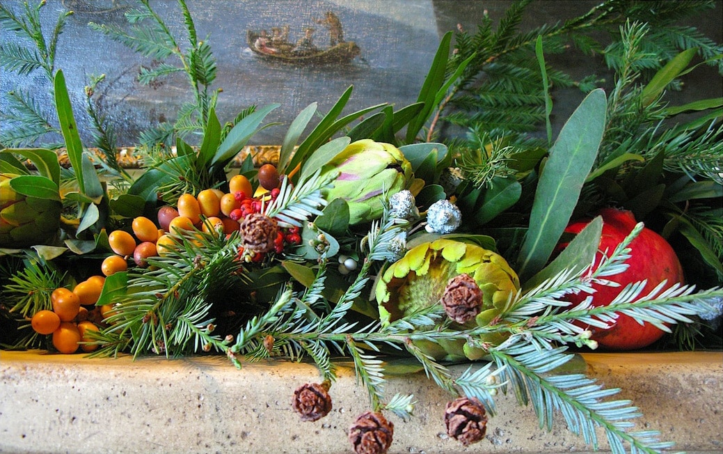 The gray concrete mantel and brown pinecones grounds the vibrant reds, golds and greens of the berries, artichokes, dates, and pomegranate.